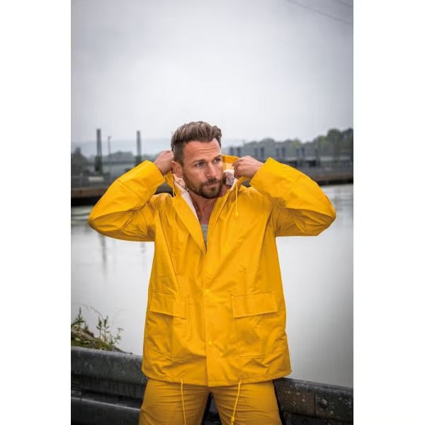 Ein Mann in einem gelben Regenmantel mit Kapuze und einer passenden gelben Regenhose steht im Außenbereich an einem Gewässer.
