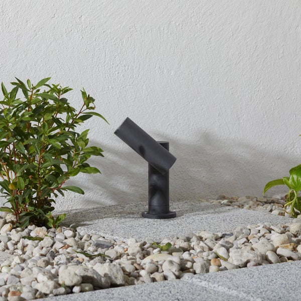 Moderner Außenstrahler in Anthrazit auf einer Steinplatte, umgeben von hellem Kies und grünen Pflanzen.