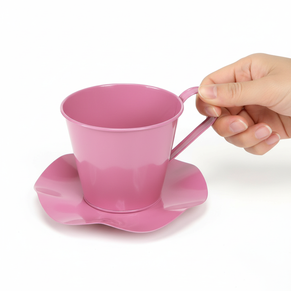 Rosafarbener Übertopf aus Metall in Form einer Tasse mit Untertasse, von einer Hand am Griff gehalten.