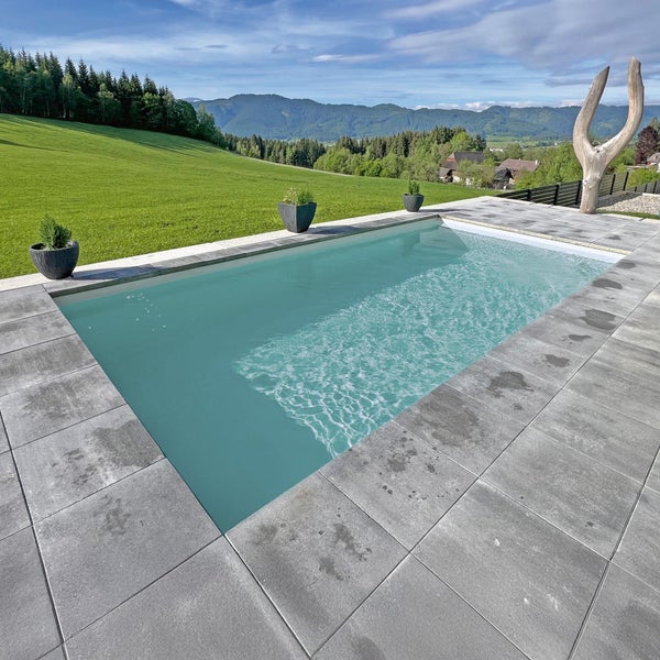Rechteckiger Außenpool eingebettet in eine Terrasse aus grauen Steinplatten, umgeben von grüner Landschaft und Bergen.
