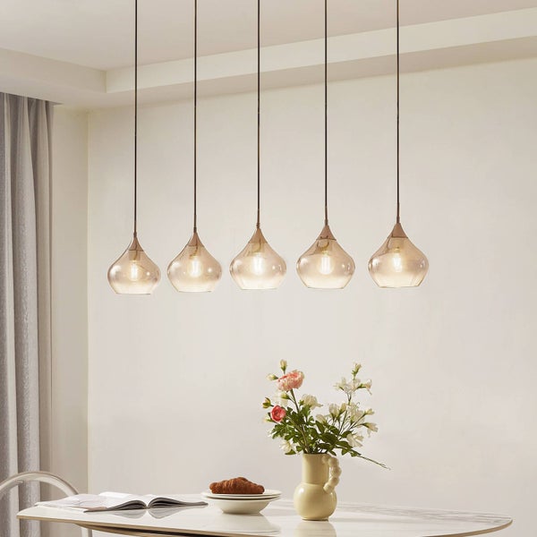 Fünf moderne Pendelleuchten mit bernsteinfarbenen Glasschirmen in einer Reihe über einem hellen Esstisch mit Blumenvase.