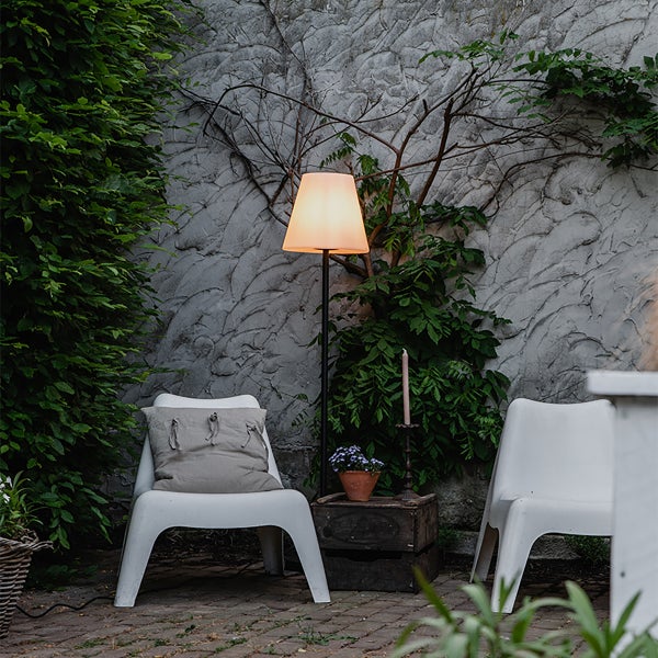 Außenstehleuchte mit Schirm in einem Garten mit weißen Sitzmöbeln, einer Holzkiste und einer strukturierten Wand mit Kletterpflanzen.
