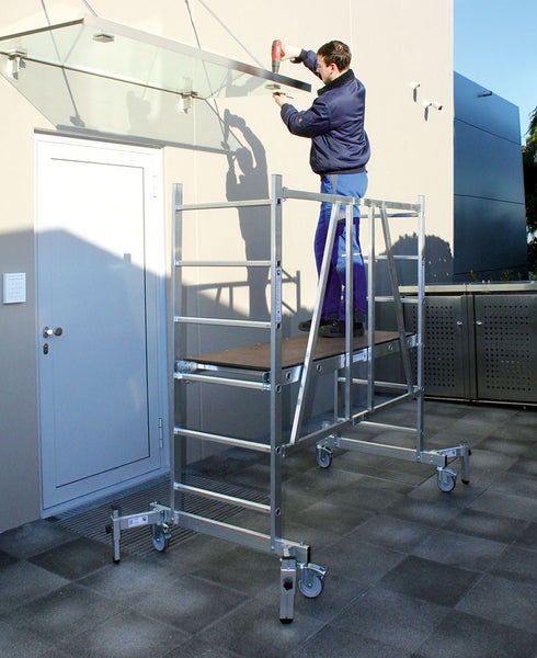 Eine Person arbeitet auf einem fahrbaren Gerüst an einer Glasüberdachung über einer Tür.