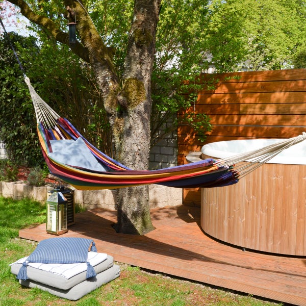 Bunt gestreifte Hängematte aus Stoff mit zwei hellblauen Kissen, befestigt an einem Baum im Garten neben einer Laterne.