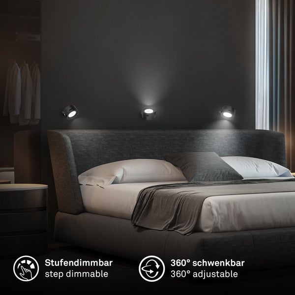 Drei kleine schwarze Wandspots über einem modernen Polsterbett in einem dunklen Schlafzimmer. Leuchten sind stufendimmbar und um 360 Grad schwenkbar.