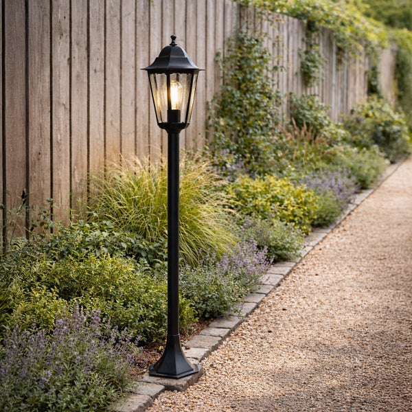Schwarze Gartenlaterne im klassischen Design an einem Kiesweg im Garten neben einem Holzzaun und Pflanzen.