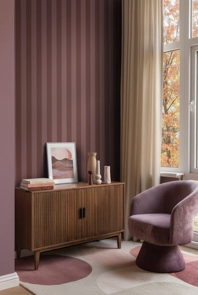 Gestreifte Tapete in Beerentönen an einer Wand in einem modern eingerichteten Wohnzimmer mit Sideboard aus Holz, Sessel und Fenster.