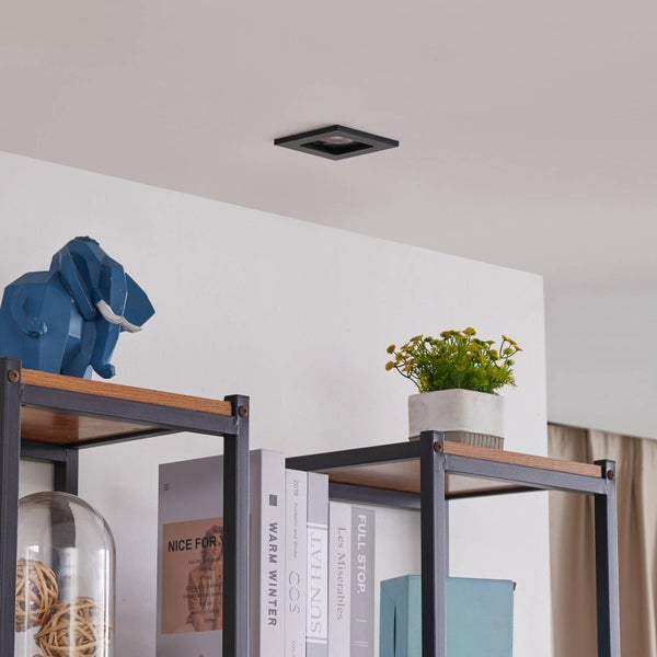 Schwarzer quadratischer Einbaustrahler in einer weißen Decke über einem modernen Metallregal mit Holzböden und Dekoration in einem hellen Raum.