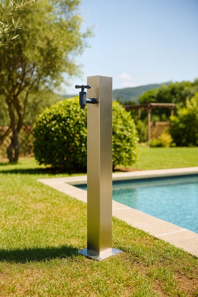 Wasserzapfsäule aus Edelstahl mit einem Wasserhahn im Garten neben einem Pool auf einer Rasenfläche.