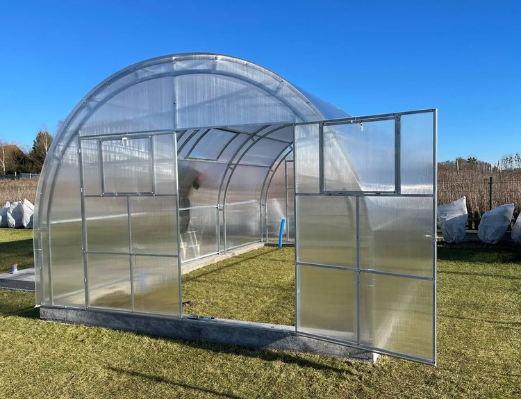 Rundbogengewächshaus mit Metallrahmen und Polycarbonat-Hohlkammerplatten auf einem Betonfundament im Garten.