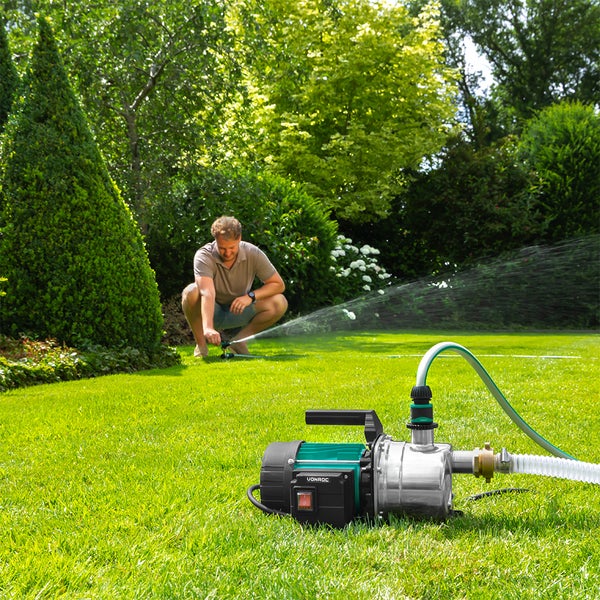 Selbstansaugende Gartenpumpe auf grünem Rasen im Garten, im Hintergrund ein Mann beim Einstellen eines Rasensprengers.