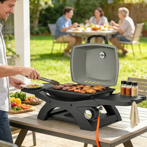 Ein Mann nutzt einen kompakten Gasgrill auf einem Tisch in einem hellen Garten. Fleisch und Meeresfrüchte liegen auf dem Grillrost.