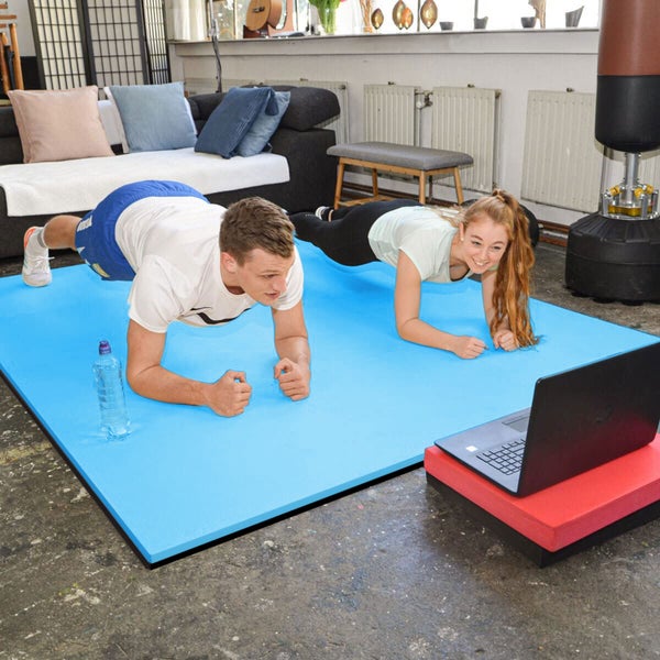 Mann und Frau führen gemeinsam ein Workout auf einer großen, texturierten Fitnessmatte in einem wohnlichen Raum mit Laptop aus.