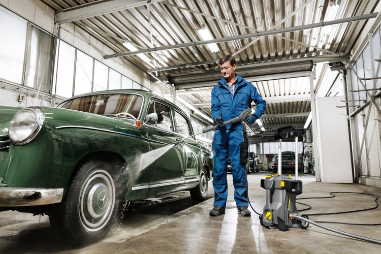 Ein Mann im blauen Overall reinigt mit einem Hochdruckreiniger einen grünen Oldtimer in einer hellen Werkstatt.