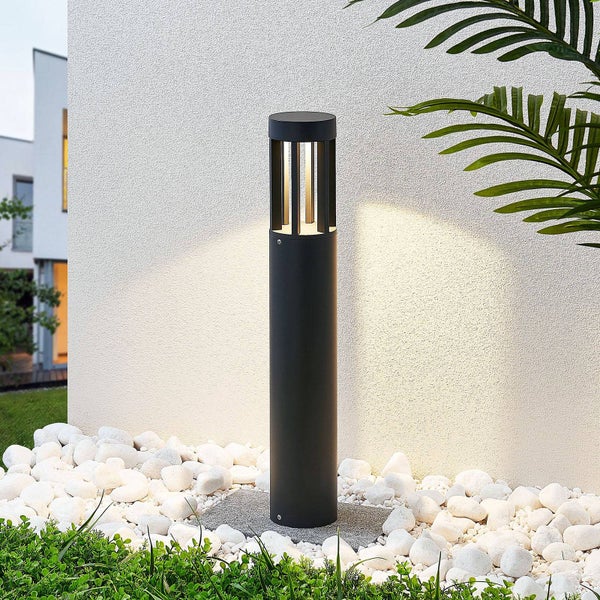 Moderne schwarze Pollerleuchte für den Außenbereich, leuchtend in einem Beet aus Kieselsteinen vor einer strukturierten Wand.