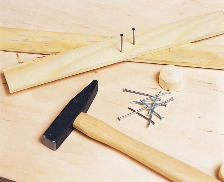 Schlosserhammer mit Holzstiel, daneben verstreute Stahlnägel und Holzleisten auf einer Werkbank.