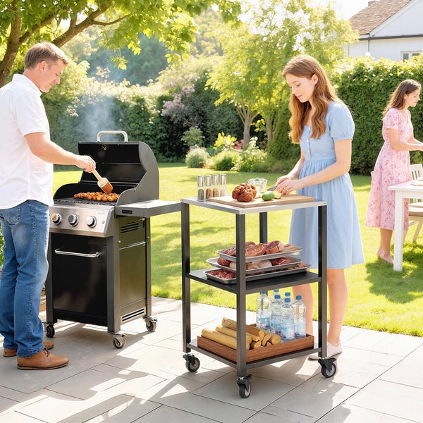 Helle Gartenszene mit schwarzem Gasgrill und dreistöckigem Servierwagen aus Metall auf einer Terrasse, Personen bereiten Essen zu.