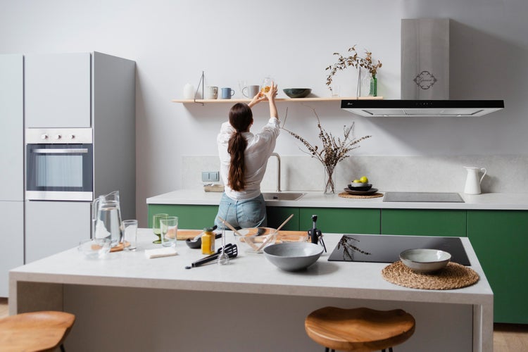 Moderne Küche mit grünen Unterschränken, weißer Arbeitsplatte, Kücheninsel und Frau, die ein Wandregal über der Spüle einräumt.