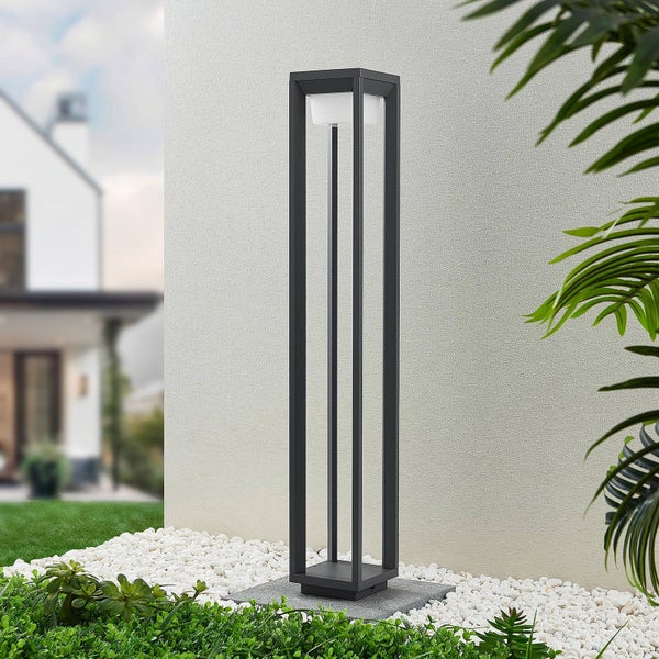 Moderne rechteckige Außen-Stehleuchte aus dunkelgrauem Metall mit weißem Diffusor, platziert in einem hellen Garten auf weißem Kies vor einer Hauswand.