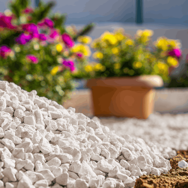 Haufen weißer Zierkies im Vordergrund einer hellen Gartenszene mit Blumen im Tontopf im Hintergrund.