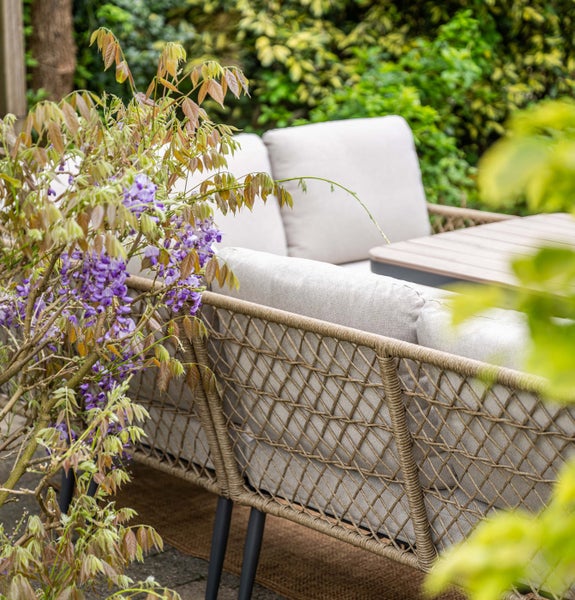 Garten-Lounge-Sofa mit Seilgeflecht-Rückenlehne und hellen Polstern in einem bepflanzten Garten.