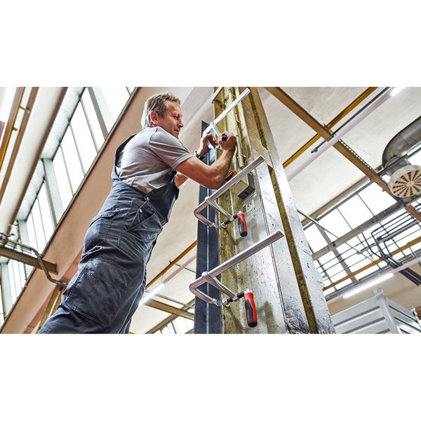 Ein Mann befestigt mit Schraubzwingen ein Werkstück an einem Pfosten in einer Werkshalle.