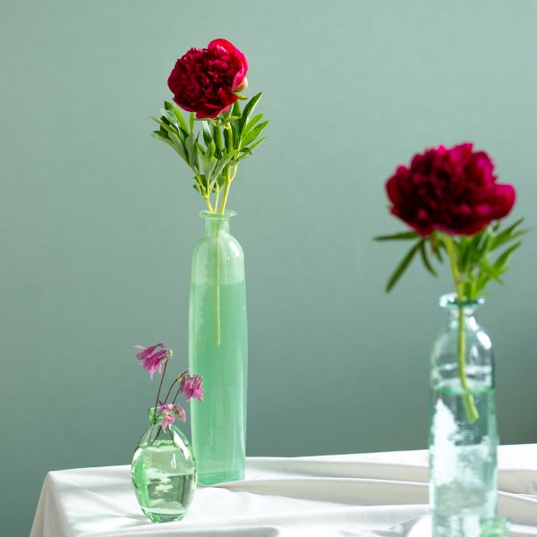 Drei Glasvasen in verschiedenen Größen mit roten Pfingstrosen und kleinen violetten Blumen auf einer weißen Tischdecke.