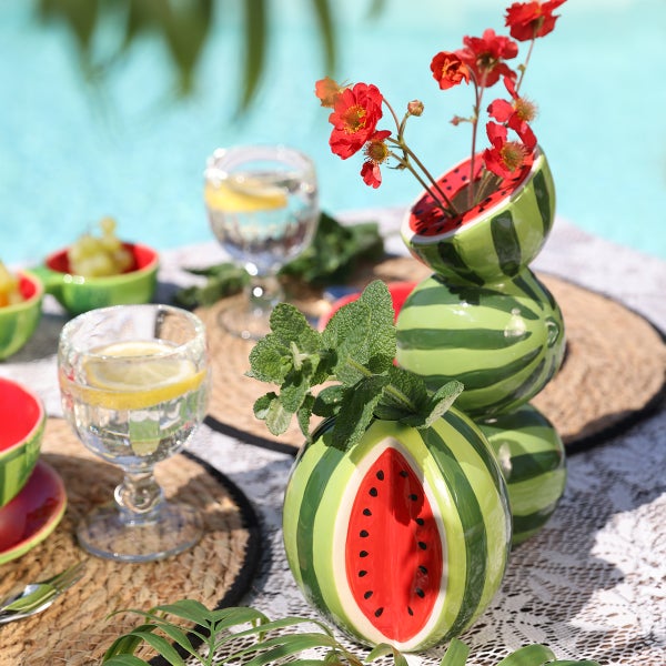 Keramikvase im Wassermelonen-Design mit roten Blumen und Minze auf einem gedeckten Tisch im Garten.