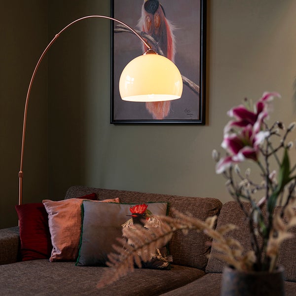 Stehlampe mit gebogenem Metallelement und kugelförmigem Lampenschirm über einem Sofa mit Kissen.