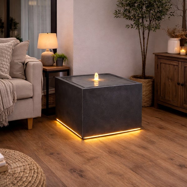 Moderner Zimmerbrunnen in Würfelform mit LED-Beleuchtung am Sockel und am Wasserstrahl in einem gemütlich eingerichteten Wohnzimmer mit Holzboden.