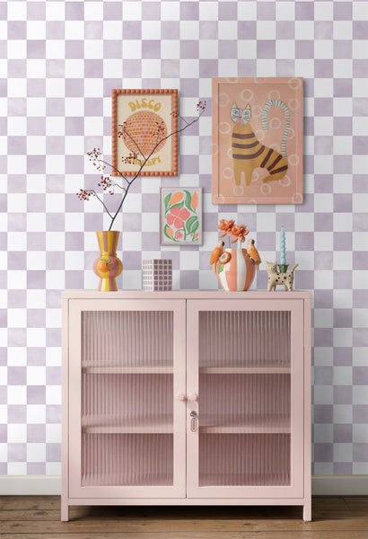 Heller Wohnraum mit lila-weiß karierter Tapete, rosa Metallschrank mit Glastüren und bunter Wanddekoration auf Holzboden.