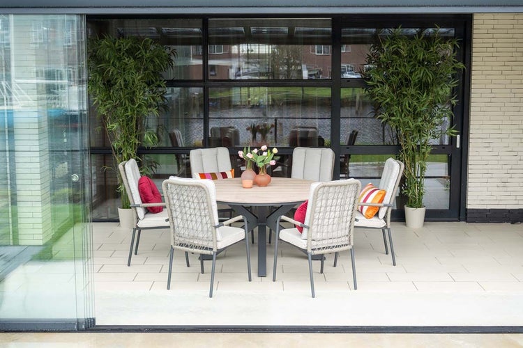 Modernes Gartenmöbel-Set mit rundem Holztisch und sechs Stühlen mit Seilgeflecht und Polstern auf einer hell gefliesten Terrasse.