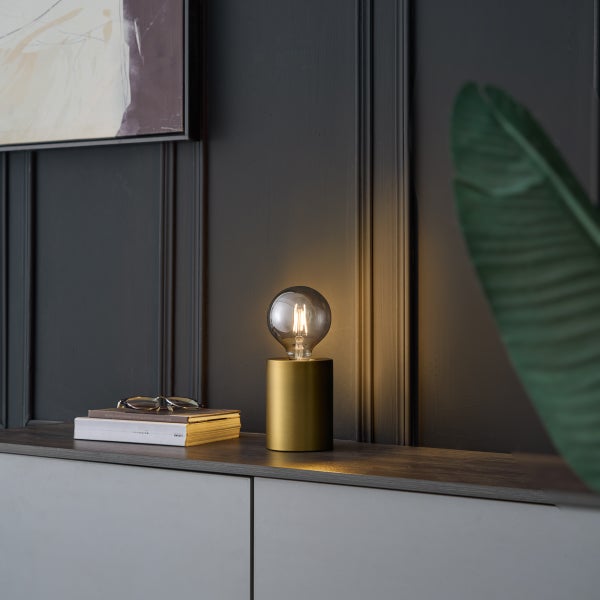 Zylindrische Tischleuchte aus Messing mit Filament-Leuchtmittel auf einem Sideboard vor einer dunklen Wand mit Holzpaneelen.