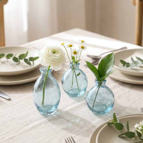 Drei kleine Vasen aus Glas auf einem gedeckten Tisch mit Blumen und Blättern als Tischdekoration.
