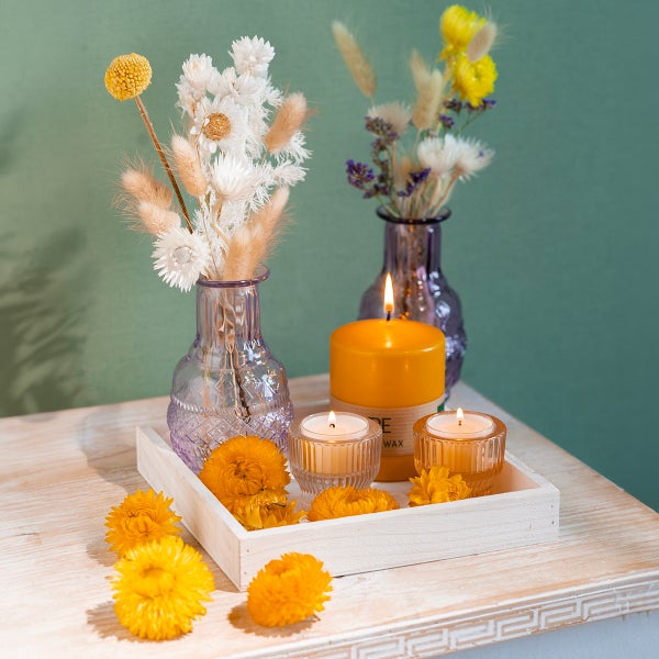 Dekorative Szene mit zwei Glasvasen mit Trockenblumen, einer Stumpenkerze und zwei Teelichtern in Glashaltern auf einem Holztablett.