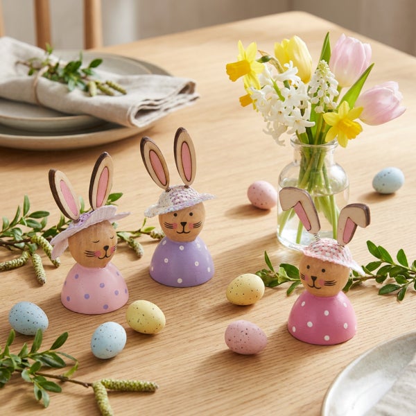 Osterdekoration mit drei hölzernen Osterhasenfiguren, bunten Ostereiern und einem kleinen Blumenstrauß aus Tulpen und Narzissen auf einem Holztisch.