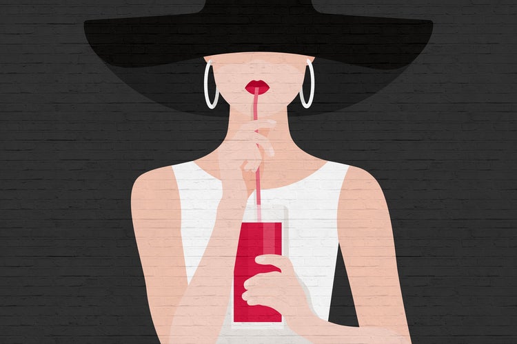 Frau mit Hut trinkt mit Strohhalm vor einer Backsteinmauer