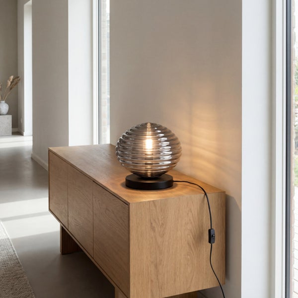 Moderne Tischleuchte mit geriffeltem Glasschirm auf einem Sideboard aus Holz in einem hellen, minimalistischen Raum.