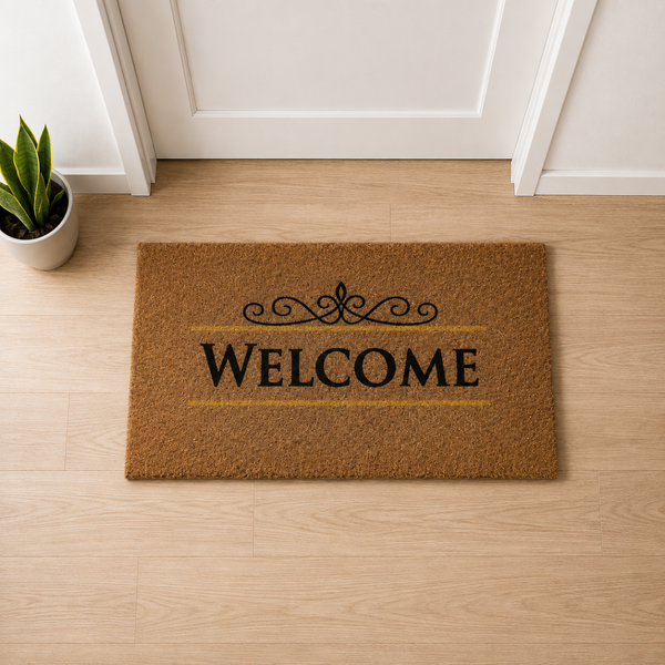 Rechteckige Fußmatte aus Kokosfasern mit dem Schriftzug WELCOME und Ornament, vor einer Tür auf einem Holzfußboden neben einer Pflanze.