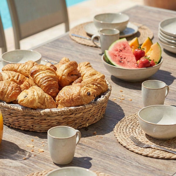 Hell gedeckter Frühstückstisch im Freien mit Keramikgeschirr, geflochtenem Korb mit Croissants, Obstschale und Platzsets auf einem Holztisch.