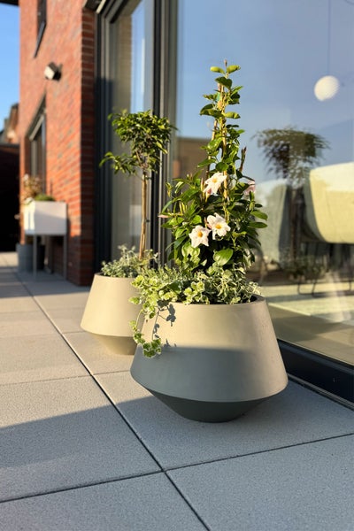 Zwei moderne, konische Pflanzkübel auf einer hellen Terrasse mit blühenden Pflanzen und einem kleinen Baum vor einer Fensterfront.
