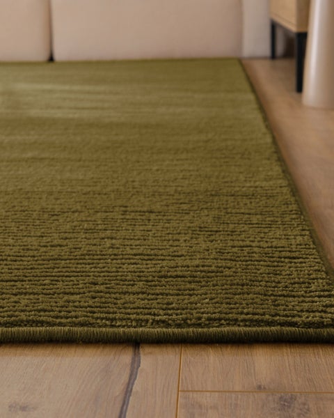 Strukturierter olivgrüner Teppich mit Rippenmuster in einem hellen Wohnzimmer auf Holzfußboden vor einem beigen Sofa.