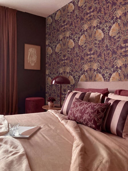 Ein Schlafzimmer mit einer gemusterten Tapete mit botanischen Motiven, einem Bett mit Kissen, einer Tischlampe und einem Hocker.