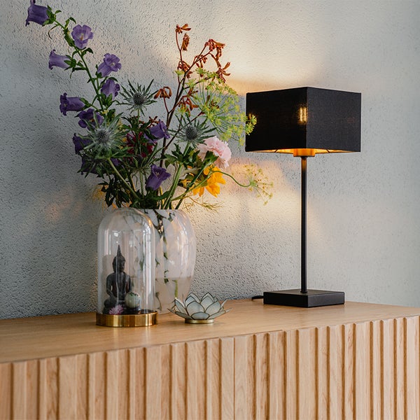 Moderne Tischleuchte mit schwarzem rechteckigem Stoffschirm auf einer Holzkommode neben einer Vase mit Blumen.