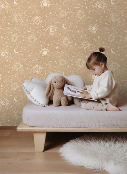Vliestapete im Kinderzimmer mit Sonnen, Monden und Sternen. Ein Kind sitzt lesend auf einem Bett vor der gemusterten Wand.