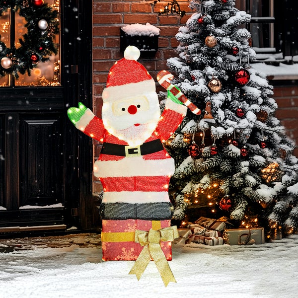 Beleuchtete Weihnachtsmann-Figur auf Geschenkbox mit Zuckerstange für den Außenbereich vor verschneitem Weihnachtsbaum und Hauseingang.