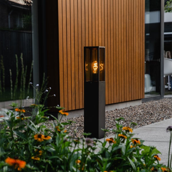 Schwarze rechteckige Wegeleuchte mit dekorativem Leuchtmittel vor einer Holzfassade im Garten.