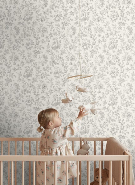 Helles Kinderzimmer mit floral gemusterter Vliestapete an der Wand, einem Holzkinderbett und einem Kind, das nach einem Mobile greift.