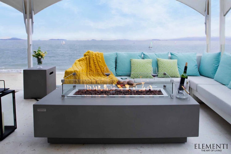 Moderner grauer Gas-Feuertisch in Rechteckform mit Glasschutz in einem hellen Outdoor-Loungebereich mit Blick auf das Meer.