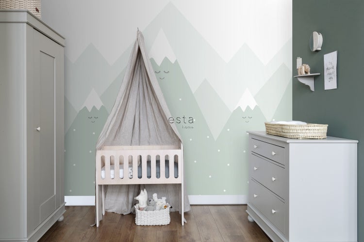 Helles Kinderzimmer mit Kinderbett, Kommode und Schrank vor einer Wand mit Bergmotiven.